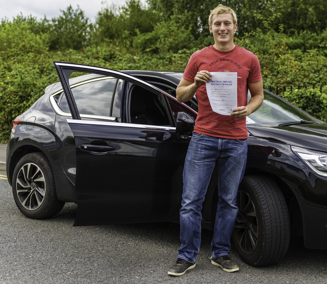 Young man standing next to black car holding blue driving test pass certificate
