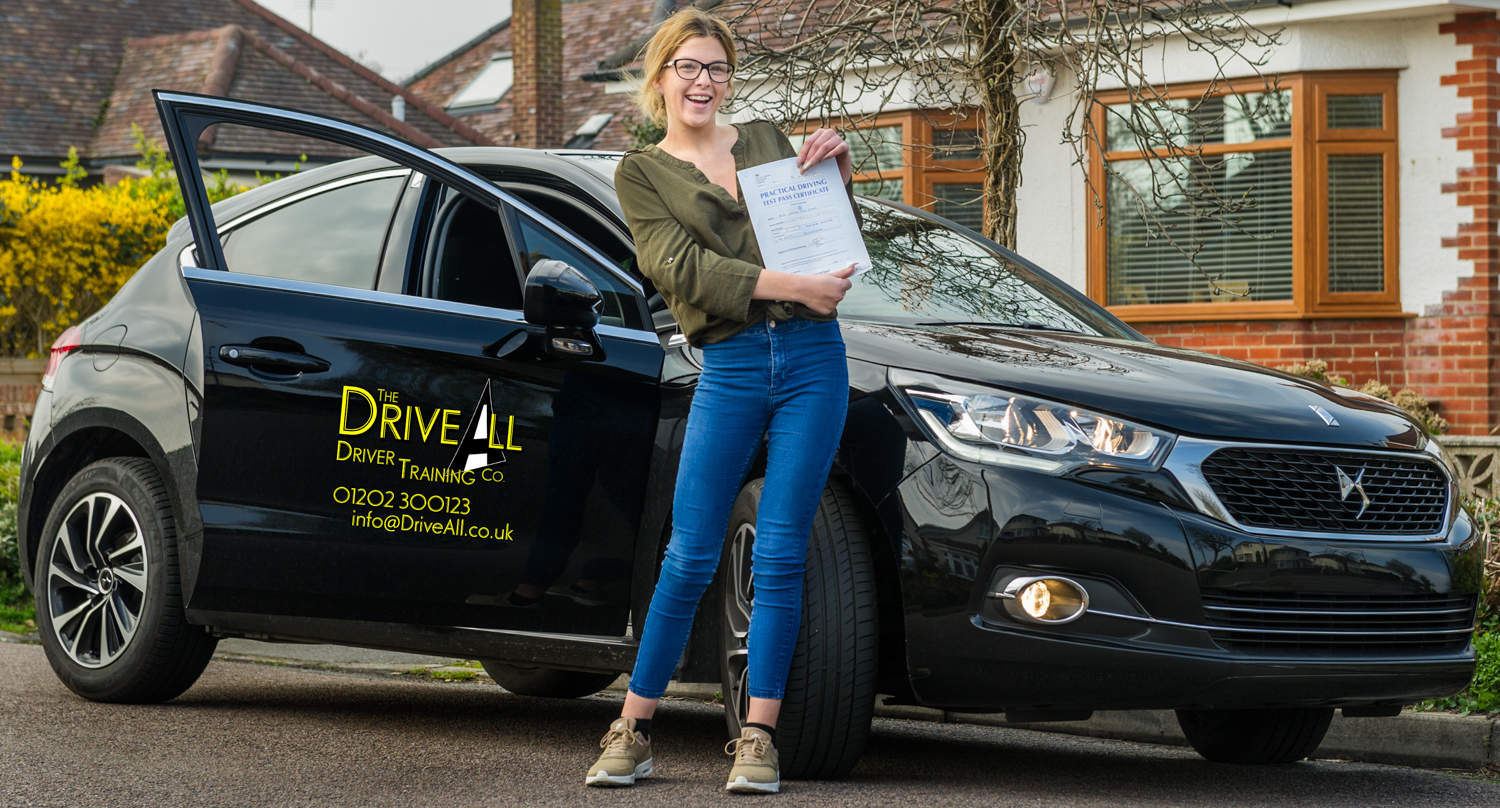 Young woman standing next to black car holding blue driving test pass certificate