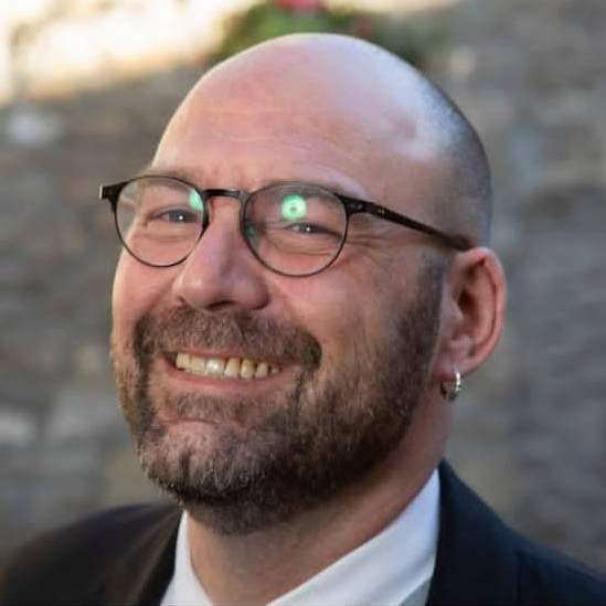 A smiling man with a short beard and shaved head, wearing black rimmed circular glasses and an round silver earing in his right ear lobe